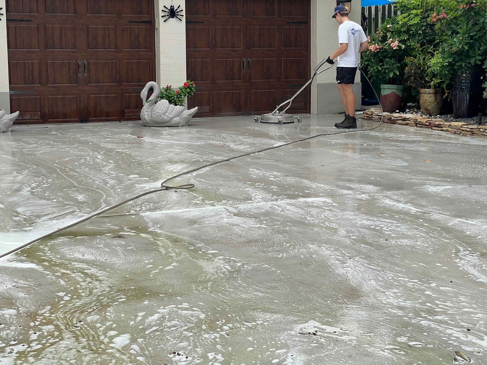 A founder in a cap and Panther Power Clean shirt running a surface cleaner across a foam-covered driveway in front of dark garage doors