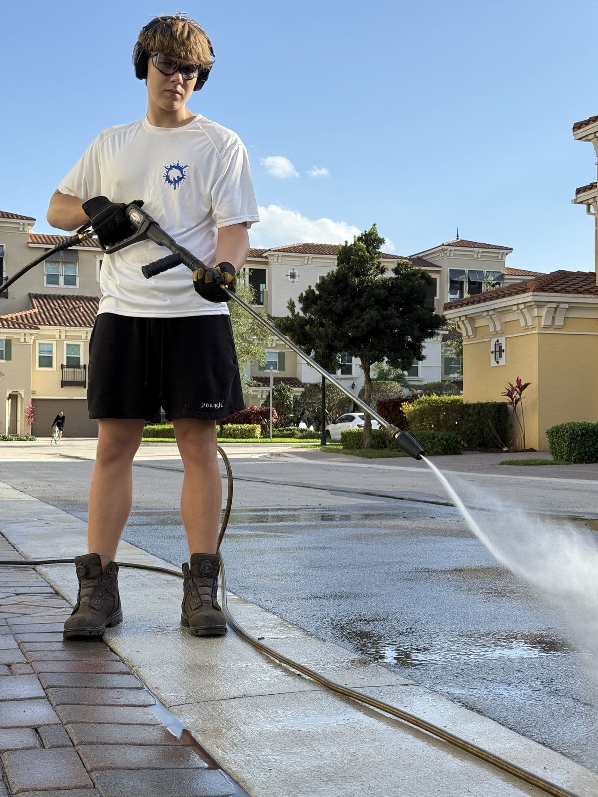 Gleb in ear protection, safety glasses and gloves, pressure-washing a driveway with a wand from a paver walkway, townhomes behind