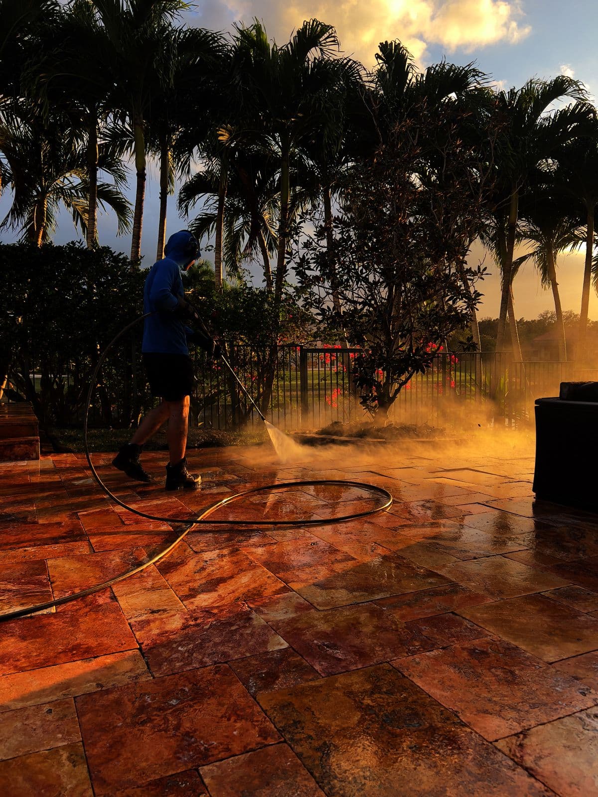 A founder silhouetted against the sunset while pressure-washing a travertine patio, the mist lit from behind