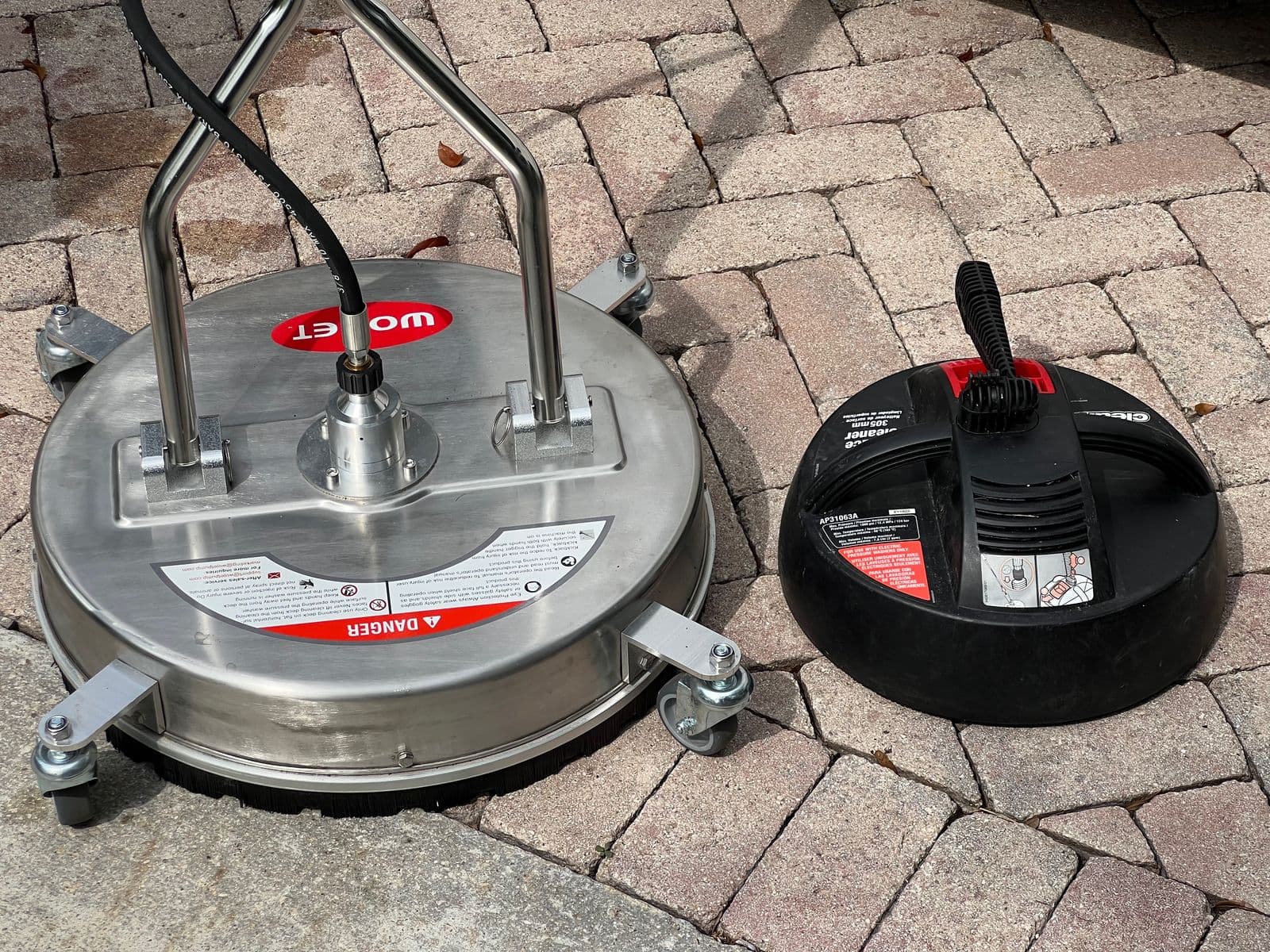 Two surface cleaners side by side on pavers: the new twenty-inch stainless one and the small black electric one from the hardware store