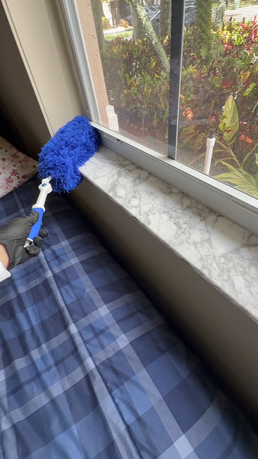 A gloved hand running a blue microfiber duster along the inside of a window frame above a marble sill, a garden outside